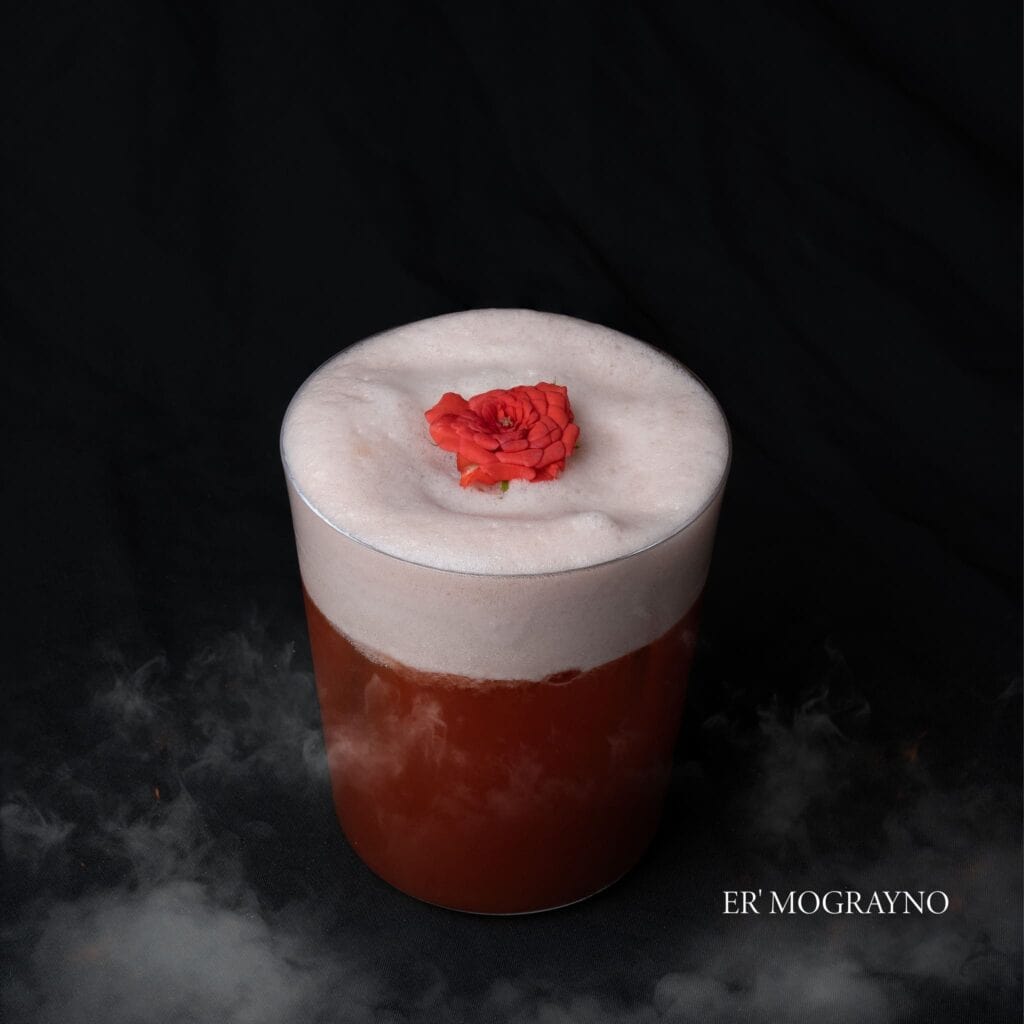 A glass of Er' Mograyno, a signature drink from Davin Cafe, topped with foam and garnished with a small red rose.