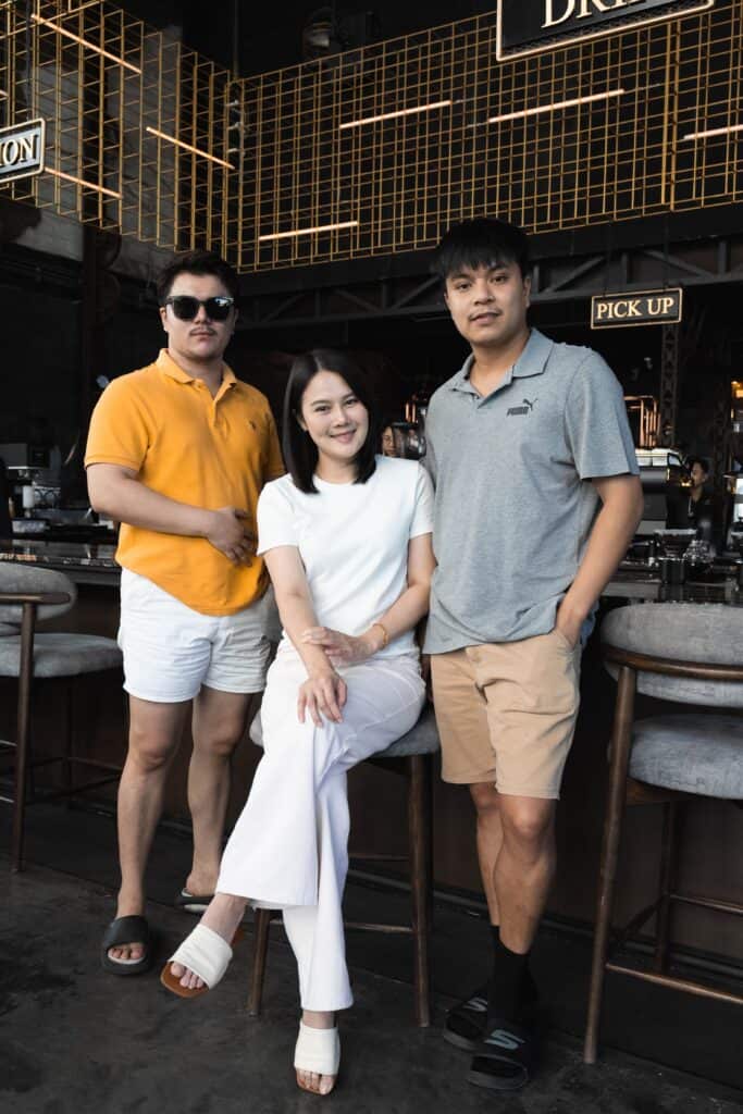 กลุ่มเพื่อนสามคนกำลังยืนและนั่งถ่ายรูปที่เคาน์เตอร์บาร์ภายในร้าน Davin Cafe.