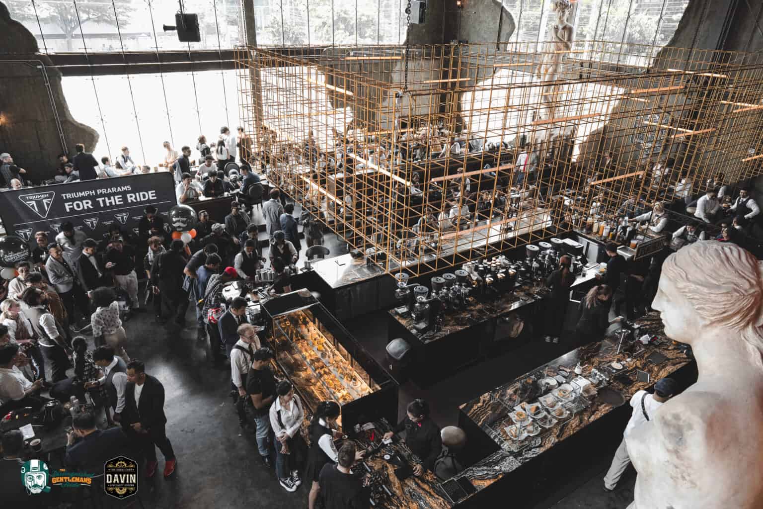 An aerial view of a Triumph Motorcycles event held at Davin Cafe, showing a large crowd of people and a unique golden grid structure.