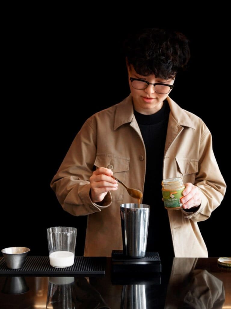 A barista wearing a tan jacket is meticulously adding ingredients to a metal shaker to prepare a craft coffee drink.