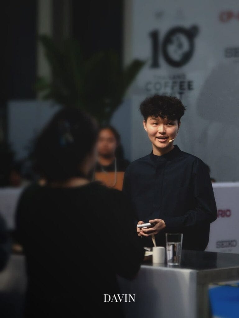 Coffee champion Jane Kawinnart smiling while speaking to an audience at a coffee event.