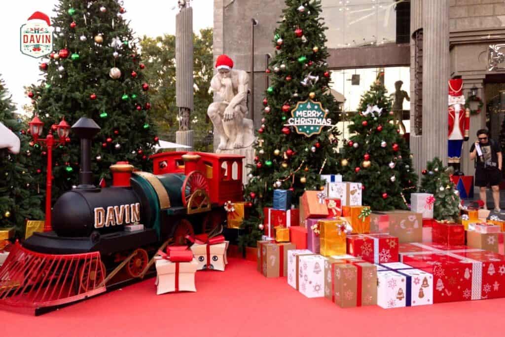 The Davin Cafe Christmas display featuring the miniature train, piles of colorful gifts, and the iconic Thinking Man statue wearing a Santa hat.