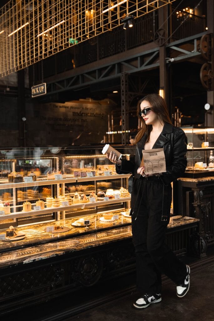A lifestyle moment at Davin Cafe, a cafe along the Ramintra Expressway, where a customer browses the dessert display while holding a coffee, highlighting the café’s premium industrial design and curated bakery experience.