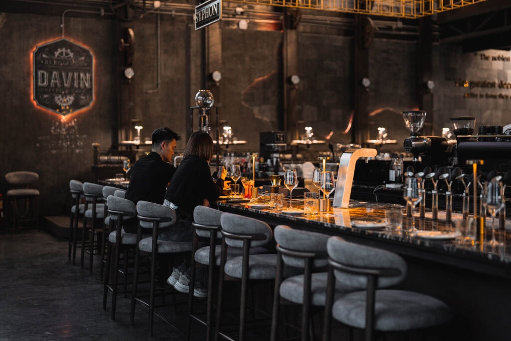 A couple enjoying an intimate dinner at the bar counter of Davin Cafe, surrounded by warm lighting and an elegant industrial interior, creating a refined evening dining experience ideal for romantic moments and relaxed conversations.