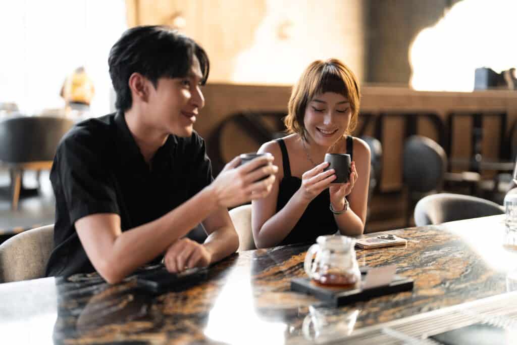 คู่ชายหญิงนั่งจิบกาแฟในคาเฟ่บรรยากาศอบอุ่นช่วงกลางวัน เหมาะสำหรับเดตแรกแบบสบาย ๆ ที่ช่วยลดความเขินและสร้างบทสนทนาอย่างเป็นธรรมชาติ