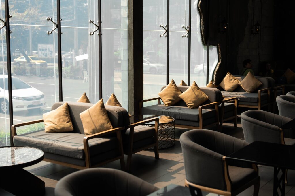 A comfortable seating area next to large floor-to-ceiling windows inside a modern coffee shop, bathed in warm natural sunlight, perfect for relaxing or casual business meetings.