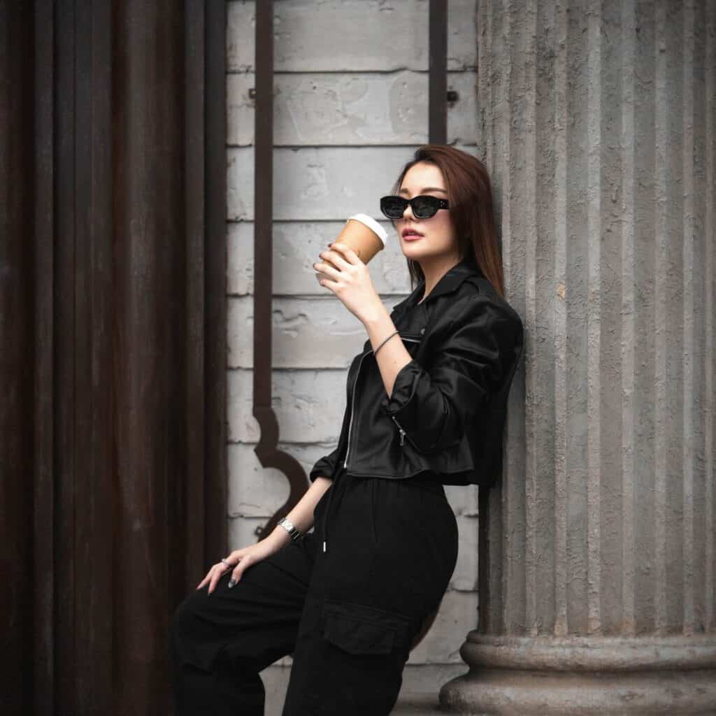 กาแฟสดในถ้วยกระดาษที่หญิงสาวใส่เสื้อแจ็คเก็ตดำและแว่นกันแดดกำลังเพลิดเพลินในบรรยากาศเมืองริมถนน.
