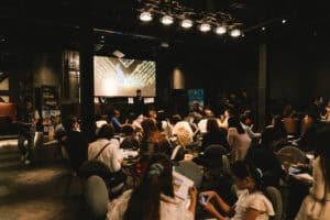 บรรยากาศการจัดงานสัมมนาทางธุรกิจที่มีผู้เข้าร่วมงานอย่างคับคั่ง โดยพื้นที่ถูกจัดสรรโต๊ะและเก้าอี้อย่างเป็นสัดส่วนเพื่อรองรับการรับฟังบรรยายบนเวทีและกิจกรรมขององค์กรได้อย่างลงตัว
