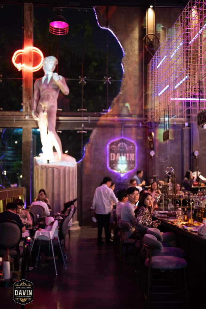 บรรยากาศภายใน Davin Cafe ช่วงค่ำที่มีผู้ร่วมงานนั่งบริเวณบาร์และพื้นที่รับประทานอาหาร ท่ามกลางแสงสี ประติมากรรม และดีไซน์ภายในที่โดดเด่น เหมาะสำหรับใช้เป็นสถานที่จัด Fan Meet ในกรุงเทพที่รองรับทั้งการพบปะแฟนคลับ กิจกรรมศิลปิน ดินเนอร์ และอีเวนต์ที่ต้องการบรรยากาศพรีเมียม