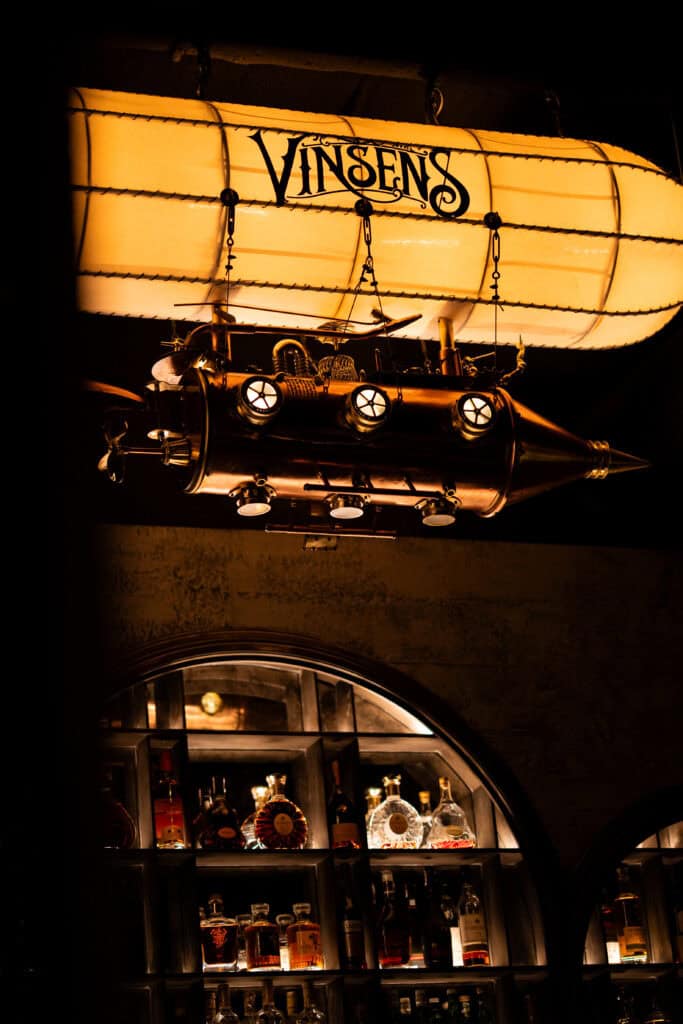 Vintage airship decor above the bar at VINSENS with amber lighting and premium bottle displays, capturing a Bangkok chill bar with a moody speakeasy atmosphere for relaxed drinks, intimate conversations, and distinctive nights out.