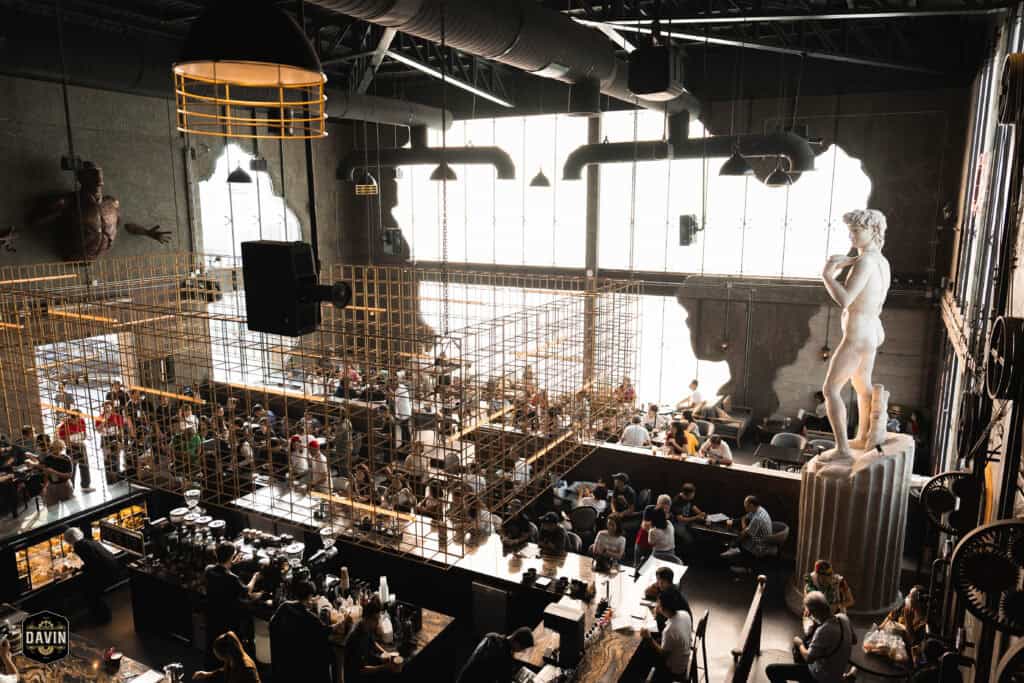 Wide interior view of Davin Cafe in Ramintra, featuring a high-ceiling European-inspired space, specialty coffee counter, spacious seating areas, and sculptural decor for a relaxed breakfast and morning cafe experience in Bangkok.