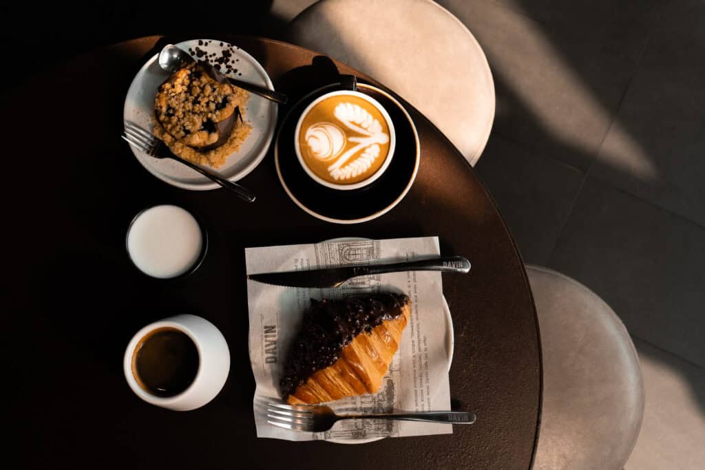 โต๊ะกาแฟและของหวานที่ Davin Cafe มีลาเต้อาร์ต เอสเพรสโซ ครัวซองต์ช็อกโกแลต และขนมอบ จัดวางในแสงธรรมชาติอบอุ่น สื่อถึงประสบการณ์ Flavor Exchange ที่เชื่อมรสชาติของกาแฟ เบเกอรี่ และโมเมนต์การชิมแบบละเมียดละไม