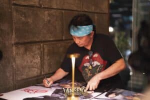 A legendary musician signing exclusive merchandise for his dedicated supporters at an intimate fan meeting venue in Bangkok.